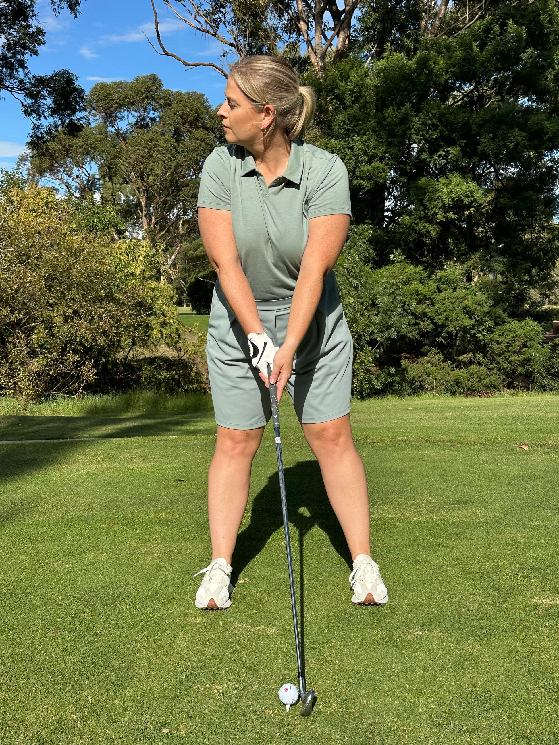 Mid-size woman wearing Fairway Green short sleeve polo and matching pleated golf shorts, preparing to swing on a sunlit course. Inclusive and performance-driven women’s golf apparel designed for focus, flexibility, and confident play.