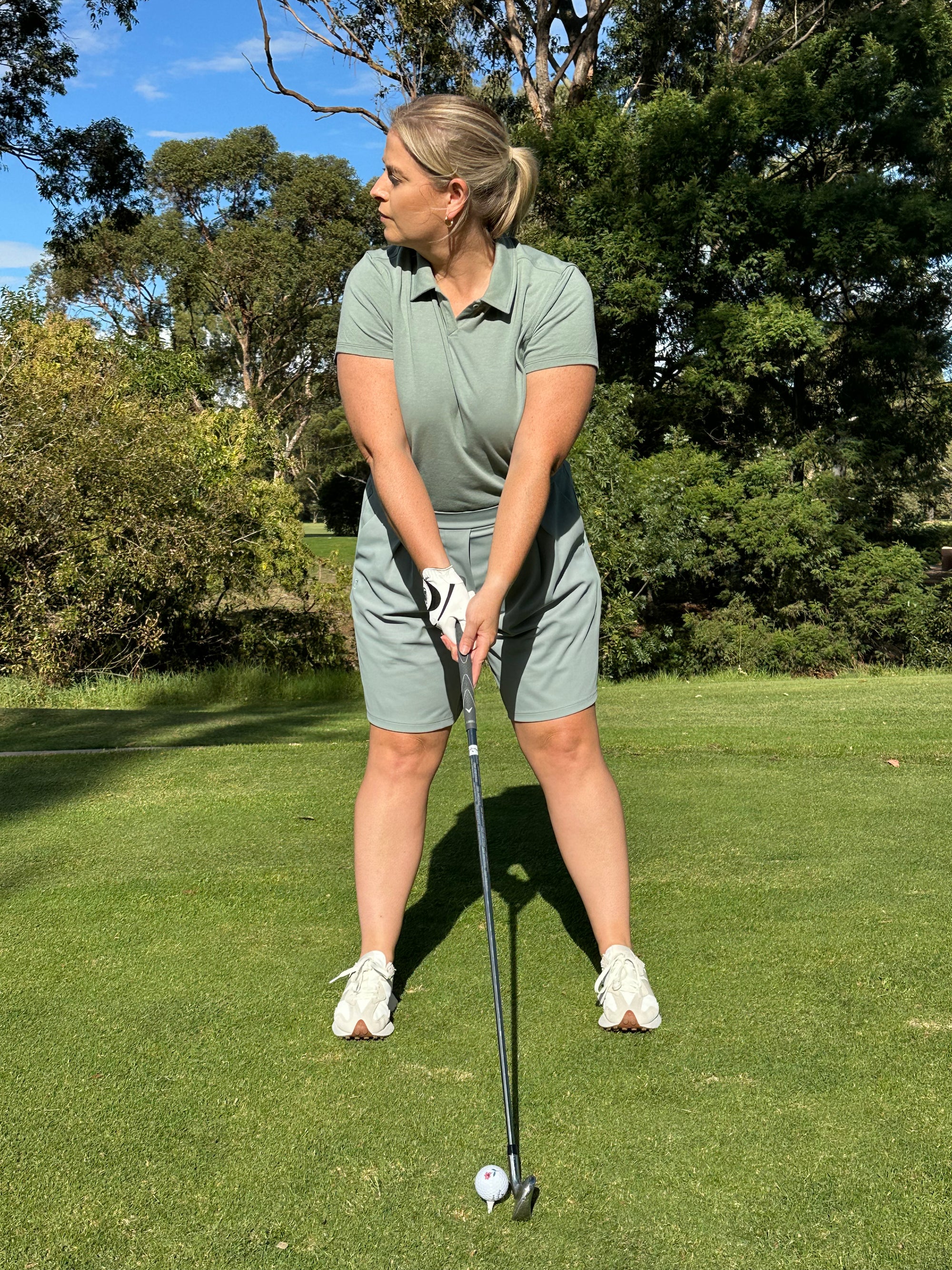 Mid-size woman wearing Fairway Green short sleeve polo and matching pleated golf shorts, preparing to swing on a sunlit course. Inclusive and performance-driven women’s golf apparel designed for focus, flexibility, and confident play.