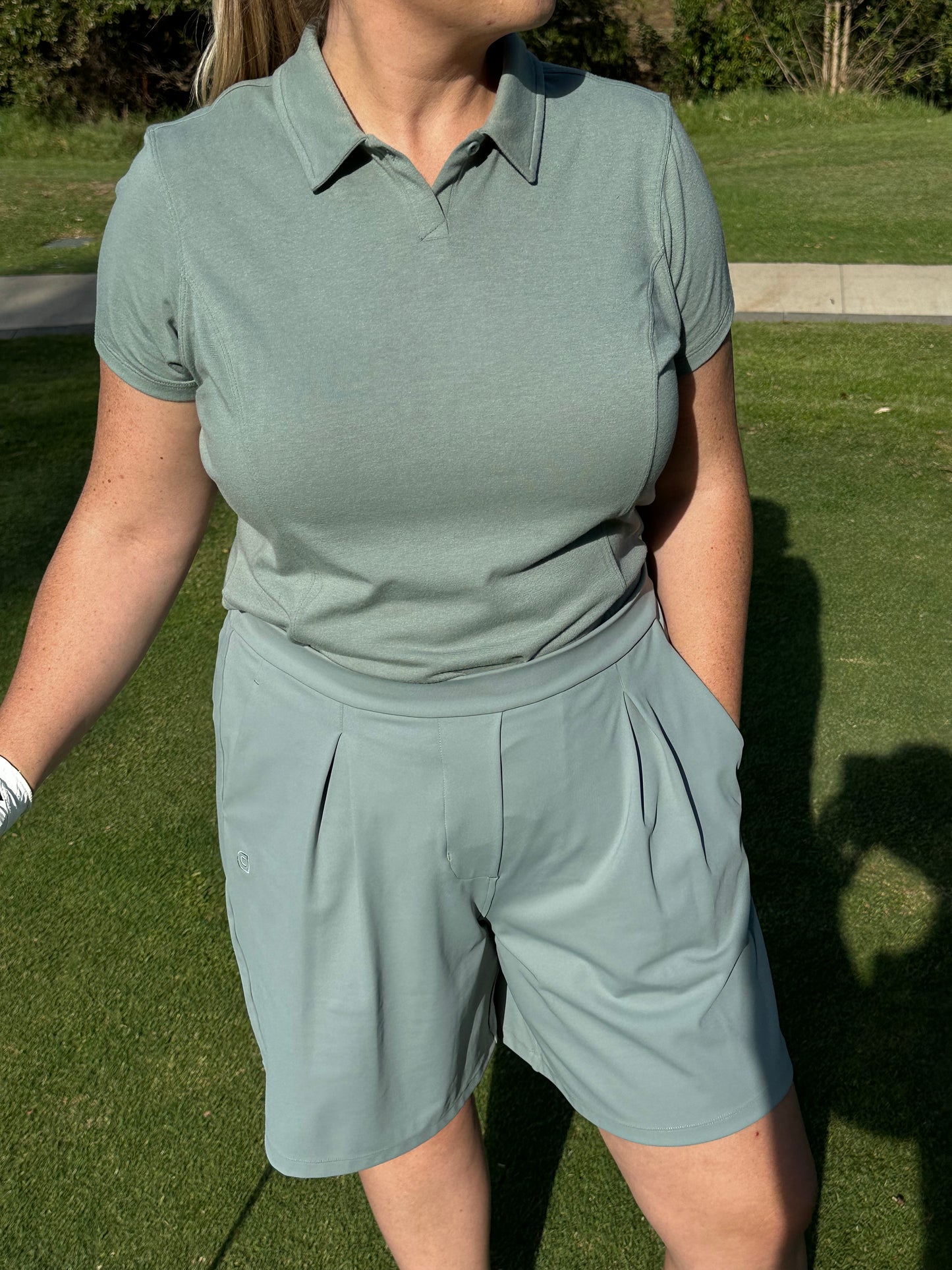 Mid-size woman wearing Fairway Green short sleeve polo and pleated golf shorts, shown from the waist up while standing on the green. Tailored and breathable women’s golf apparel offering comfort, coverage, and polished athletic style.