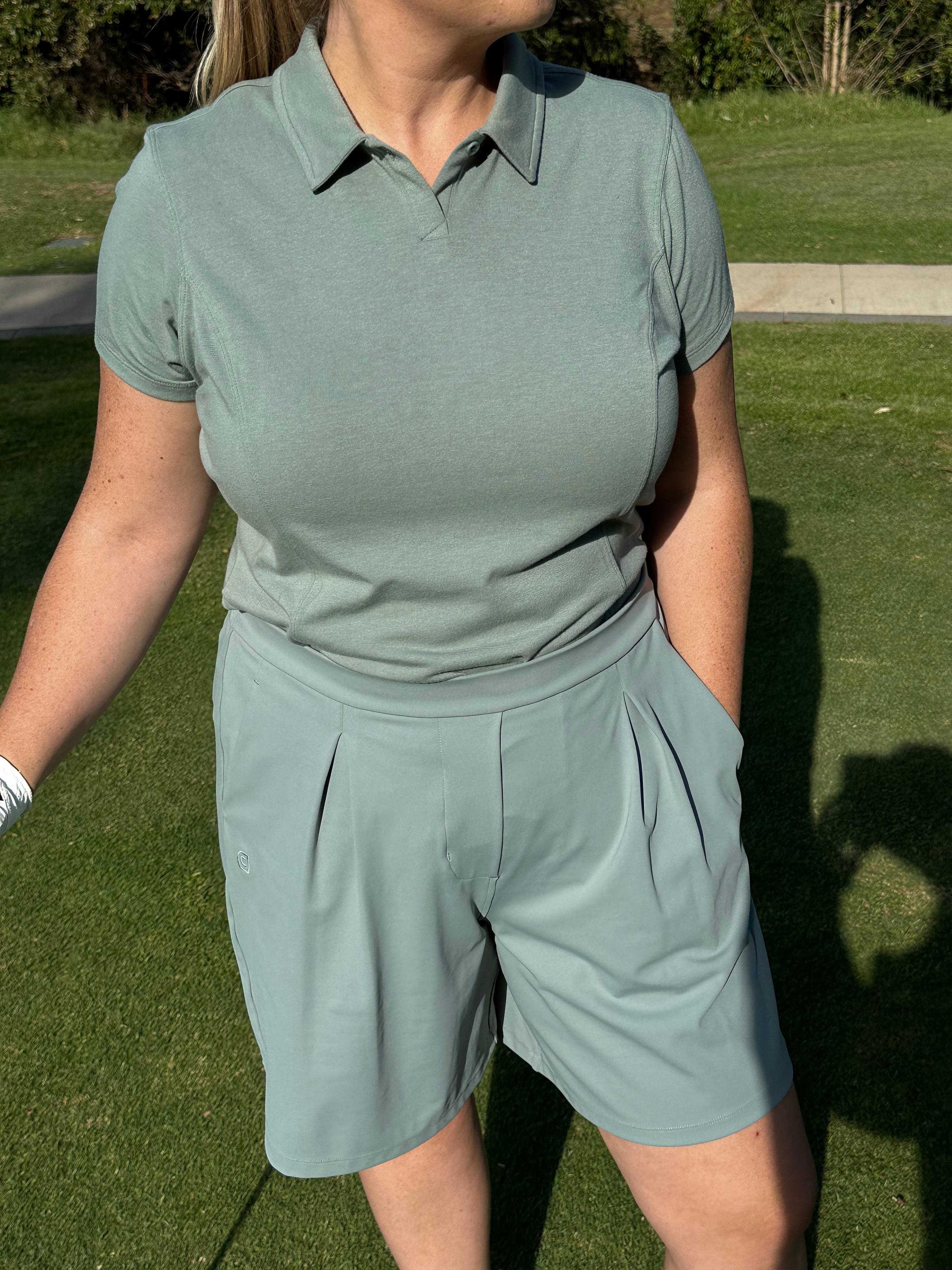 Mid-size woman wearing Fairway Green short sleeve polo and pleated golf shorts, shown from the waist up while standing on the green. Tailored and breathable women’s golf apparel offering comfort, coverage, and polished athletic style.
