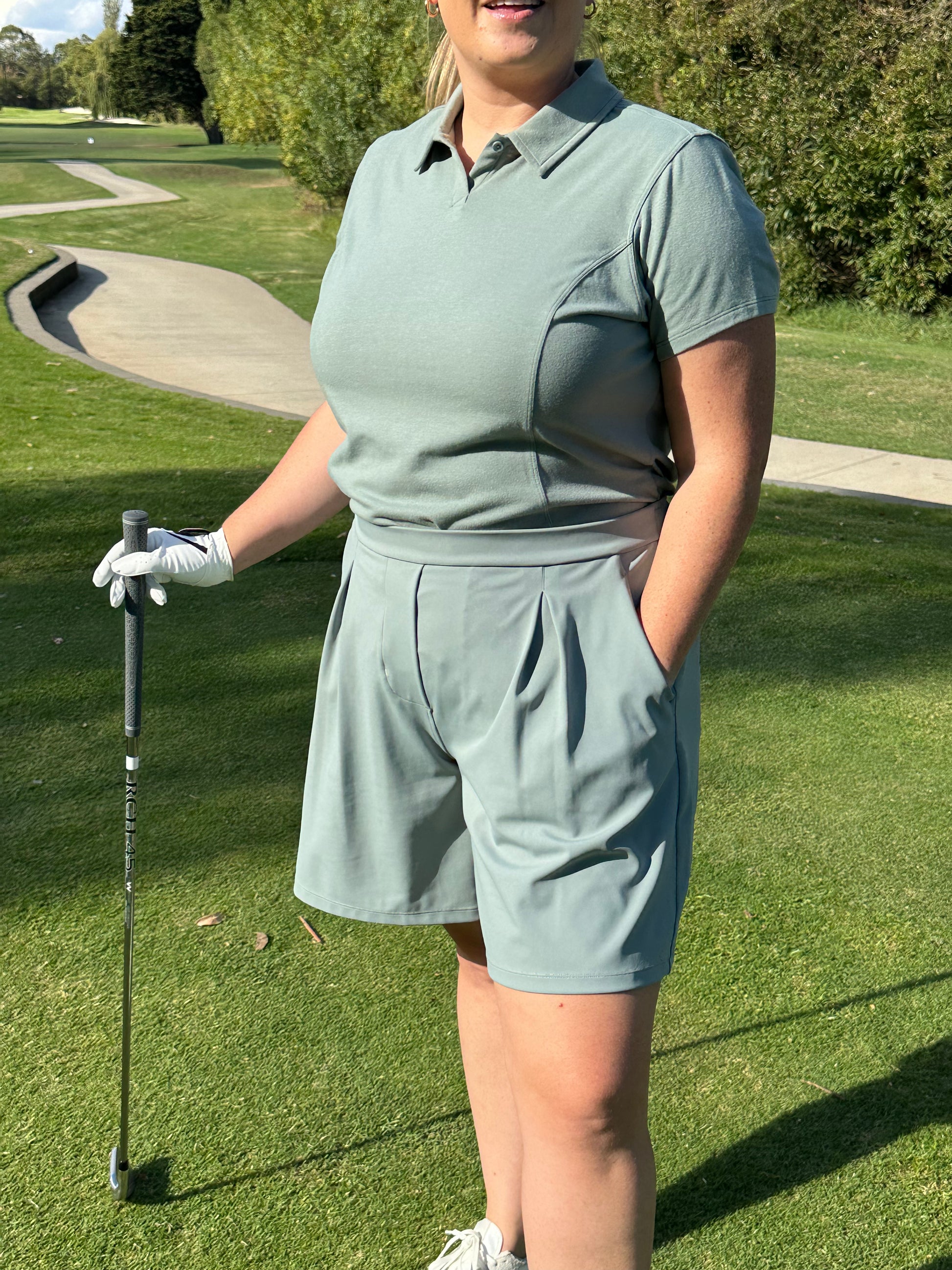 Mid-size woman wearing Fairway Green short sleeve polo and matching pleated golf shorts, standing confidently with one hand in pocket and holding a golf club on the green. Functional and flattering women’s golf apparel tailored for movement and modern athletic elegance.