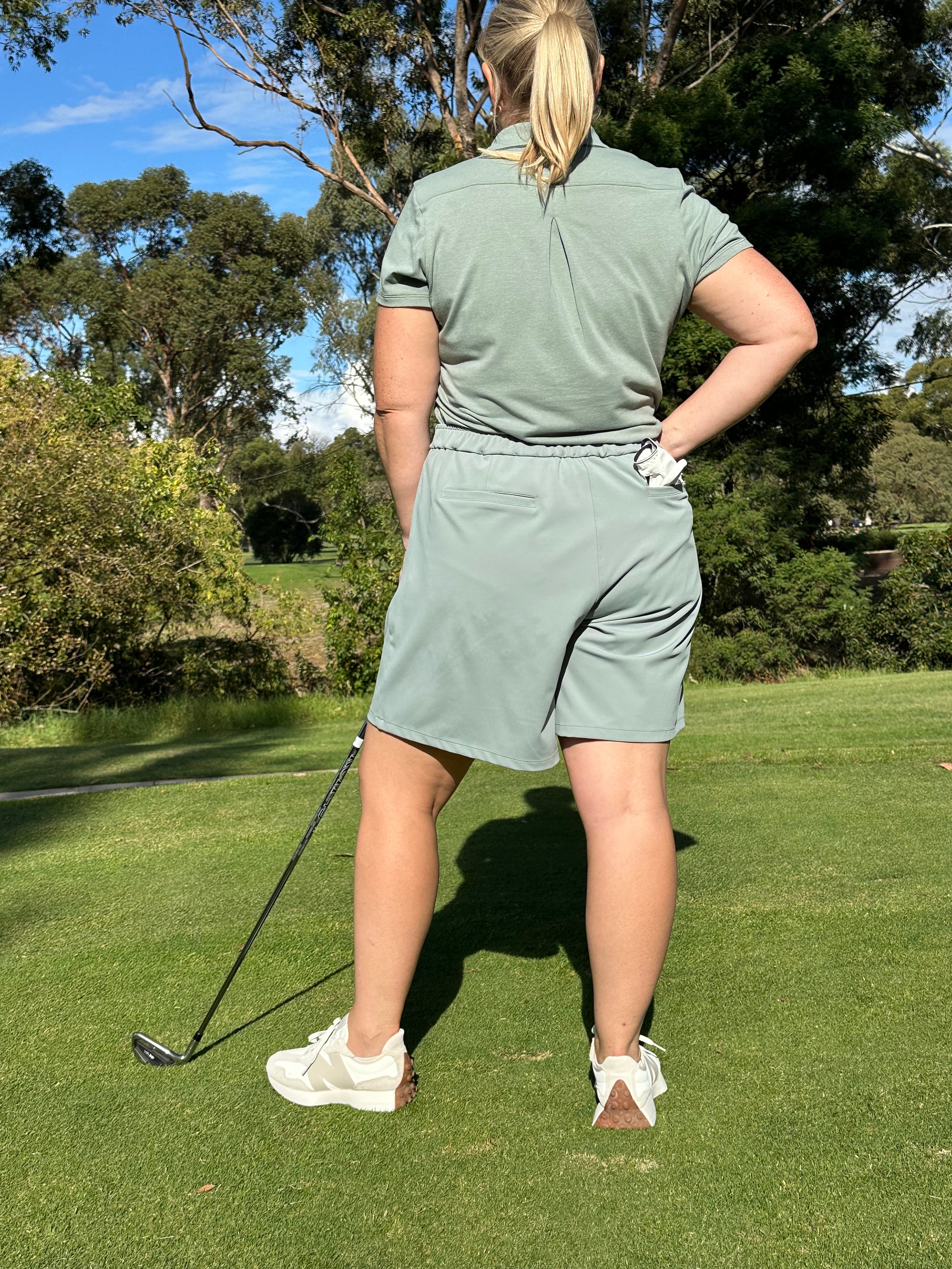Mid-sized woman wearing Fairway Green short sleeve polo and Fairway Green pleated golf shorts, standing with a golf club on a lush course, back view. Elegant and functional women’s golf apparel designed for confident and active players.