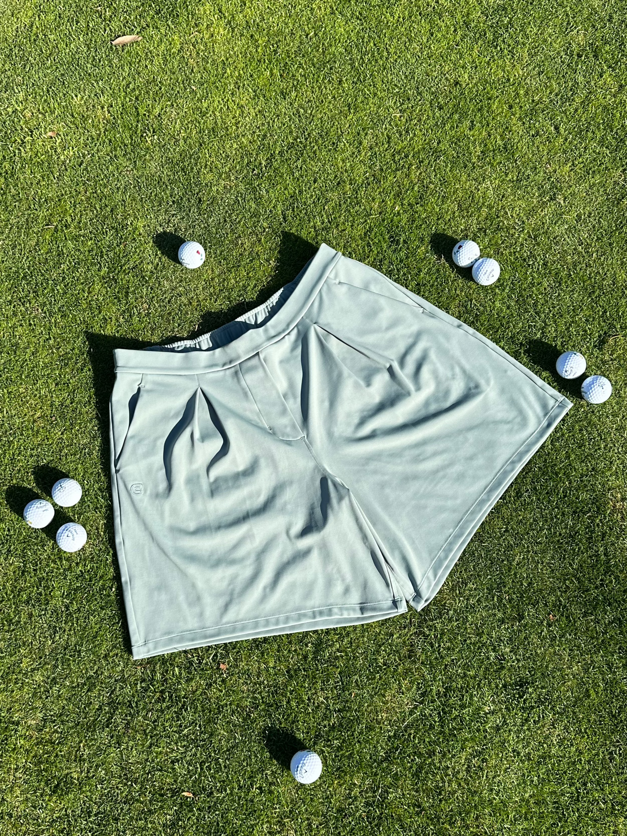 Flat lay of Fairway Green pleated golf shorts with elastic waistband, displayed on green grass surrounded by scattered white golf balls. Sophisticated and sporty women’s golf apparel designed for performance and comfort on the course.